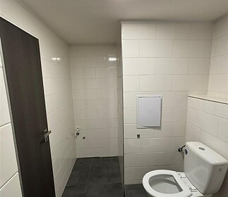 Prázdná koupelna s bílými kachlovými obklady, tmavou dlažbou a moderní toaletou, připravená k rekonstrukci.