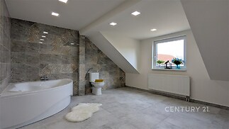 Prostorná koupelna s moderní dlažbou, rohovou vanou a velkým oknem, nabízí světlý interiér s podkrovním designem.