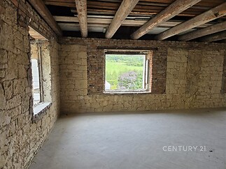 Hlavní obývací prostor s rustikální kamennou zdí, velkými okny a dřevěným stropem nabízí rekonstrukční potenciál a výhled do přírody.