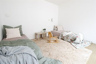 Dětský pokoj s dřevěnou podlahou, světlým kobercem a bílými stěnami, obsahující dvě postele. Minimalistický styl a útulná atmosféra.