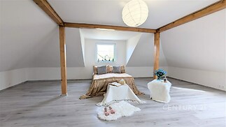 Podkrovní ložnice s moderními dřevěnými trámy, světlou parketovou podlahou a velkým oknem poskytujícím dostatek přirozeného světla.