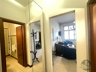 Úzká předsíň s dlažbou, velké zrcadlo a vestavěná skříň, vedle moderního obývacího pokoje s velkými okny a pohodlnými křesly v rekonstruovaném bytě.