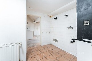 Prázdná kuchyň s bílými dlaždicemi, dlažbou na podlaze a přístupem do vedlejší místnosti, vhodná pro rekonstrukci a modernizaci prostoru.