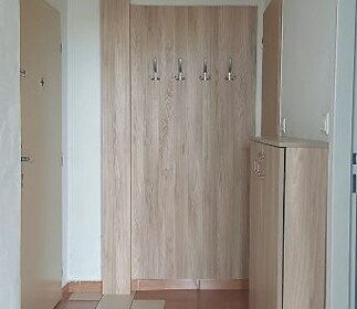 Chodba s dlažbou a dřevěným obložením, vestavěná skříň a moderní věšák na kabáty, světlý prostor vhodný pro úložné řešení.