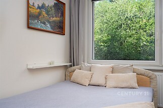 Útulná ložnice s velkým oknem, které poskytuje výhled do zeleně. Moderní styl s světlým povlečením a příjemným obrazem na stěně.