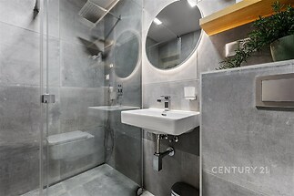 Moderní koupelna s šedou dlažbou, sprchovým koutem s posuvnými dveřmi, kulatým zrcadlem a závěsným umyvadlem v minimalistickém stylu.
