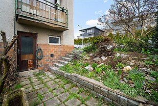 Venkovní terasa s dlažbou a udržovanou zahradou, nabízí přístup k domu. Terasa je obklopena zelení a kamennými prvky, vhodná pro relaxaci.