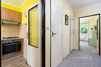 Úzká kuchyň s parketovou podlahou a dřevěnými skříňkami, přilehlá chodba s moderním designem a světlými stěnami.