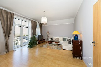 Světlý obývací pokoj s parketovou podlahou, velká okna a moderní světlý design, televize na stolku, beige sedací souprava, dekorační lampa.