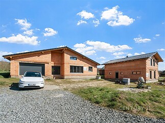 Novostavba domu s garáží a druhou budovou. Velká okna, moderní architektura, neomítnuté cihlové zdi, klidná lokalita poblíž přírody.