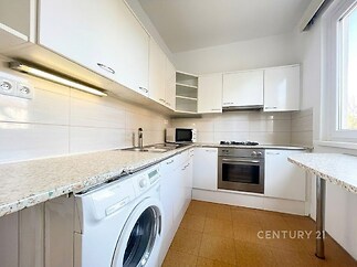 Moderní kuchyň s bílými skříňkami, vestavěnou troubou a pračkou, dlažba na podlaze, velké okno zajišťující dostatek světla.