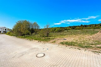 Stavební pozemek s dlážděnou příjezdovou cestou, okolní zelení a výhledem na lesní krajinu, ideální pro moderní rodinný dům.