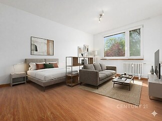 Prostorná ložnice s parketovou podlahou a velkými okny, moderní nábytek včetně šedé pohovky a manželské postele, minimalistický styl.