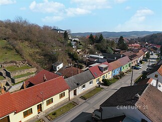 Pohled na klidnou rezidenční ulici s barevnými domy a červenými střechami, obklopenou zelení a kopcovitou krajinou, ideální pro rodinné bydlení.