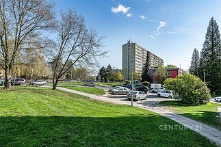 Prázdný veřejný prostor s trávníkem a chodníkem, panelový bytový dům v pozadí, stromy lemující ulici, klidná rezidenční oblast.