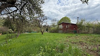 Zahrada s travnatým pozemkem obklopující dřevěnou chatu, stromy poskytující stín, klidná lokalita ideální pro rekreaci a relaxaci.