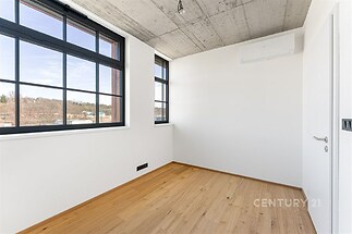 Světlý pokoj s velkými okny, moderní dřevěná podlaha a betonový strop, ideální pro rekonstruovaný industriální styl.