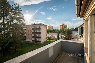 Balkón s výhledem na panelové domy, obklopený zelení. Betonová podlaha a prostorný výhled dotvářejí klidnou atmosféru moderního bydlení.