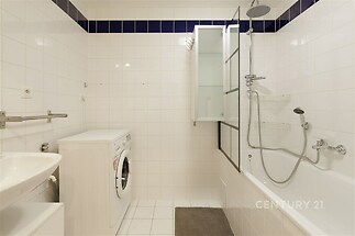 Koupelna s bílými kachličkami, vanou a sprchovou hlavicí. Moderní instalace, dlažba a praktická pračka zajišťují pohodlí.