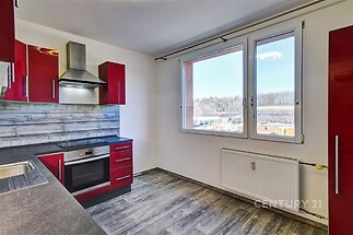 Moderní kuchyň s červenými skříňkami, dřevěným obkladem a velkými okny, parketová podlaha vytváří světlý a prostorný vzhled.