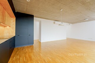Prostorný obývací pokoj s parketovou podlahou, moderní dřevěné a černé skříně, betonový strop a velká okna zajišťující světlý interiér.