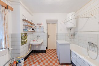 Koupelna s dlaždicovou podlahou a bílými dlaždicemi na stěnách, vybavená vanou a umyvadlem. V rohu je umístěn bojler na ohřev vody.