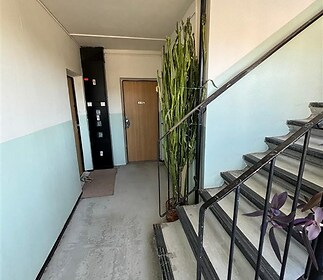 Společná chodba bytového domu, betonová podlaha, kovové zábradlí, vstupní dveře s dřevěným povrchem, klidný prostor.