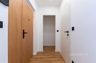 Úzká chodba s dřevěnou podlahou, bílými stěnami, moderními dveřmi a černými kovovými doplňky, vytváří čistý a světlý prostor.