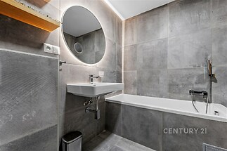 Moderní koupelna s šedými dlaždicemi, velkým kulatým zrcadlem a vestavěnou vanou. Stylové osvětlení a elegantní umyvadlo.