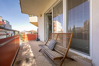 Prostorný balkon s dlažbou, dřevěnou lavičkou a velkými francouzskými okny, moderní městský výhled a slunná expozice.