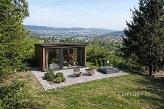 Malý zahradní domek s velkými okny a moderním designem, obklopený zelení, nabízí panoramatický výhled na krajinu a město