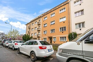 Čtyřpatrový bytový dům s oranžovou fasádou a bílými okny, nacházející se v klidné rezidenční čtvrti s parkovacími místy před domem.