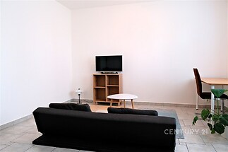 Světlý obývací pokoj s dlažbou, minimalistický design, moderní nábytek, televize na dřevěné polici, prostor působí vzdušně a čistě.