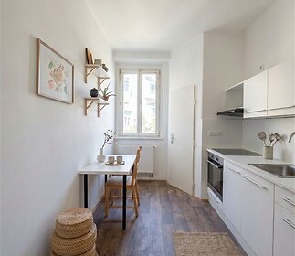 Kompaktní kuchyň s moderními spotřebiči, dřevěnou podlahou a světlými skříňkami, velké okno poskytuje dostatek přirozeného světla.