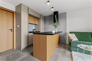 Moderní kuchyň s dřevěnou linkou, parketovou podlahou a vestavěnými spotřebiči, vedle níž je světlý obývací pokoj se zelenou pohovkou.
