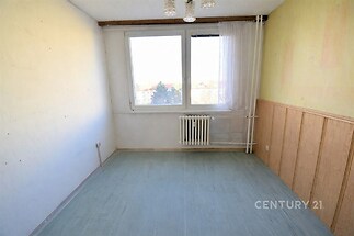 Jednoduchý pokoj s velkým oknem, parketovou podlahou a světlými stěnami, vhodný pro rekonstrukci.