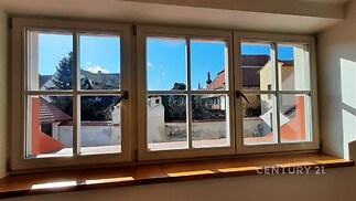 Velká okna s dřevěnými rámy, světlá místnost s výhledem na historickou architekturu a klidnou zahradu.