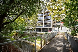 Moderní bytový dům s velkými okny a balkony, obklopen zelení, v klidné lokalitě u potoka, ideální pro příjemné bydlení ve městě.