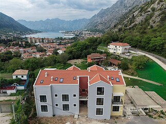 Moderní bytový dům s červenou střechou a velkými okny, nacházející se v malebné oblasti s výhledem na hory a nedaleké jezero.