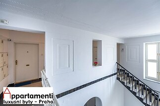 Společná chodba bytového domu, světlá se dlažbou a dřevěným zábradlím, zrekonstruovaná s bílými stěnami a moderními svítidly.