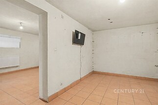 Prázdná místnost s dlažbou a bílými stěnami, vhodná k rekonstrukci, světlá díky oknu, obsahuje radiátor pro vytápění.