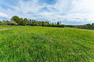 Prázdný pozemek s velkou travnatou plochou, obklopený přírodou a zalesněným horizontem, ideální pro výstavbu nebo zemědělské využití.