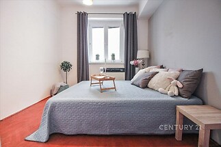Prostorná ložnice s parketovou podlahou, velké okno zajišťující světlo, moderní nábytek a dekorace, světlé stěny a závěsy.
