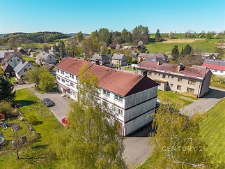Vícepatrový bytový dům s červenou střechou a bílou fasádou, umístěný v klidné rezidenční oblasti, obklopený zelení a zahradami.