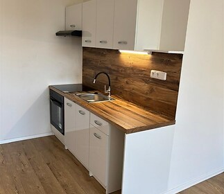 Moderní kuchyň s dřevěnou pracovní deskou a parketovou podlahou, vybavená světlými skříňkami a vestavěnou troubou.