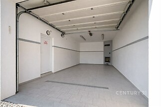 Garáž s keramickou dlažbou, elektrickými vraty a bílými stěnami. Prostorná a dobře osvětlená, vhodná pro parkování i skladování.