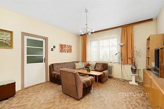 Světlý obývací pokoj s hnědou sedací soupravou, kobercem a velkými okny, zdobený béžovými stěnami a dekoračními prvky v retro stylu.