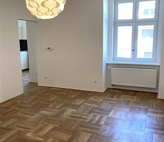 Světlý obývací pokoj s parketovou podlahou, velkými okny a moderním osvětlením, ideální pro komfortní bydlení.