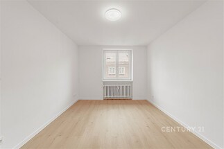 Světlý pokoj s parketovou podlahou, velkým oknem a moderní bílou malbou, ideální pro rekonstrukce nebo jako prázdné plátno pro kreativní design.