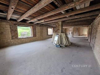 Prostorná rekonstruovaná místnost s odhalenými dřevěnými trámy, betonovou podlahou a velkými okny, ideální pro moderní úpravu.